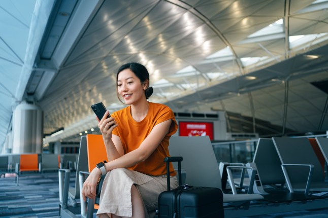Young Asian woman with suitcase using smartphone while waiting for her flight at airport terminal. Asian businesswoman on business travel. Lifestyle and technology. Travel and vacation concept